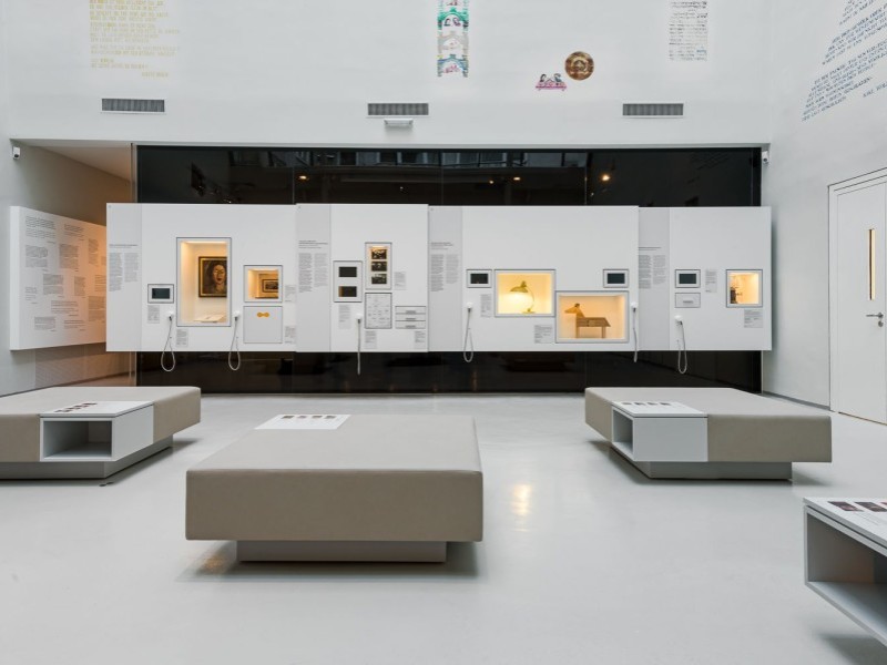 Moderne Museumsausstellung mit beleuchteten Vitrinen, Texttafeln und Sitzbänken in hellem Raum