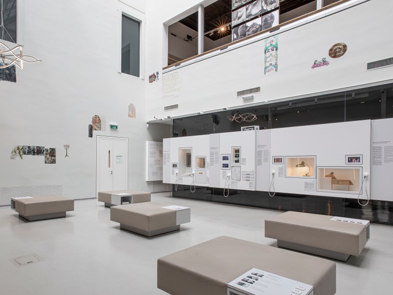Heller Ausstellungsraum mit weißen Wänden im Jüdischen Museum Wien, weiße Sitzbänke, beleuchtete Vitrinen mit Objekten an der rechten Wand und Doseninstallation links