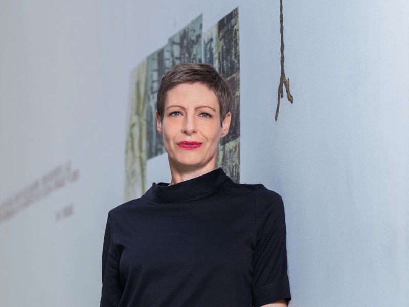 Barbara Staudinger steht vor einer weißen Wand mit einer Menora und Fotos, trägt ein schwarzes Oberteil mit halblangen Ärmeln und schwarze Hose