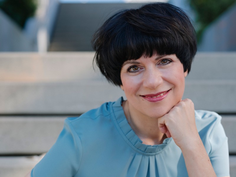 Frau mit kurzem dunklem Haar in blauem Oberteil sitzt auf einer Treppe und stützt ihr Kinn auf die rechte Faust