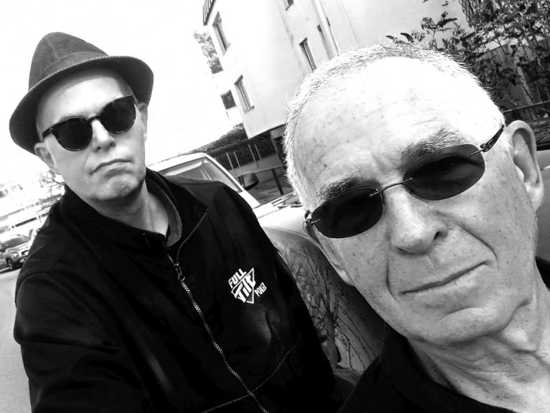 Zwei Männer stehen im Freien vor einem mehrstöckigen Wohngebäude, einer trägt einen Hut und eine Jacke mit einem sichtbaren Logo.