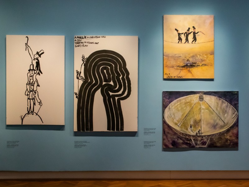 Fünf Gemälde an einer blauen Wand in einem Museum, darunter schwarz-weiße Linienzeichnungen und farbige Bilder mit menschlichen Figuren und abstrakten Formen.