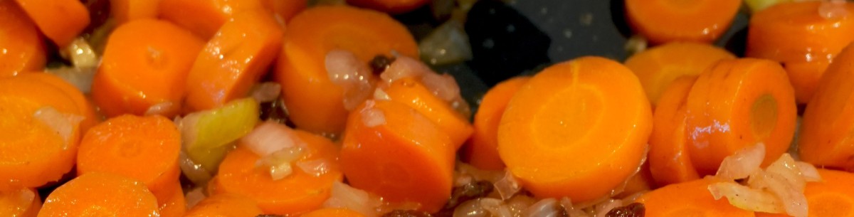 : In einer Pfanne liegen geschnittene Karotten, Zwiebeln und Sellerie. Die Karotten sind orange und rund, einige sind mit Rosinen vermischt.