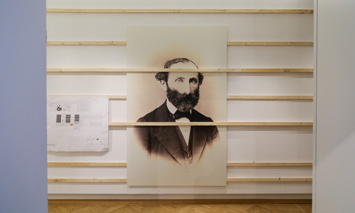 Schwarz-weiß-Porträt eines bärtigen Mannes in Anzug und Fliege, befestigt an einer weißen Wand mit horizontalen Holzlatten