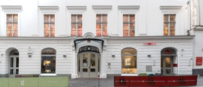 Fassade des Jüdischen Museums Wien: Weiße klassizistische Fassade mit Eingangsportal und fünf sichtbaren Fenstern, vor dem Gebäude zwei Sitzbereiche mit Glasabschirmungen