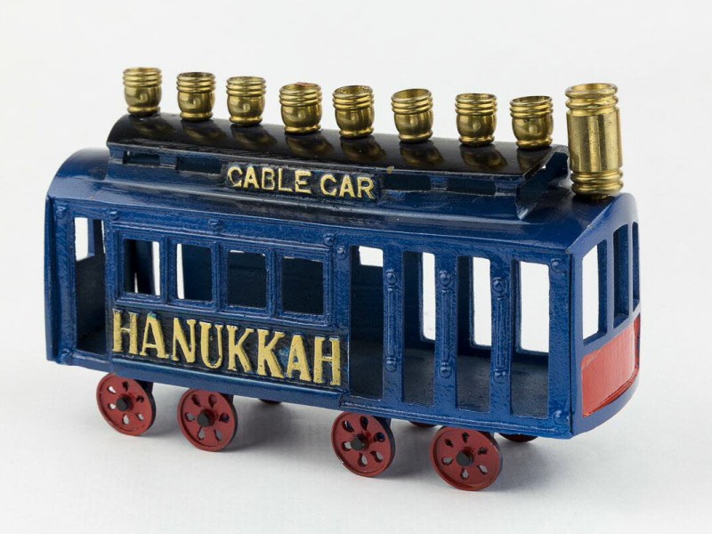 Blaues Modell einer Straßenbahn mit roten Rädern und goldfarbenen Chanukka-Kerzen auf dem Dach, Schriftzug 'CABLE CAR' und 'HANUKKAH'.