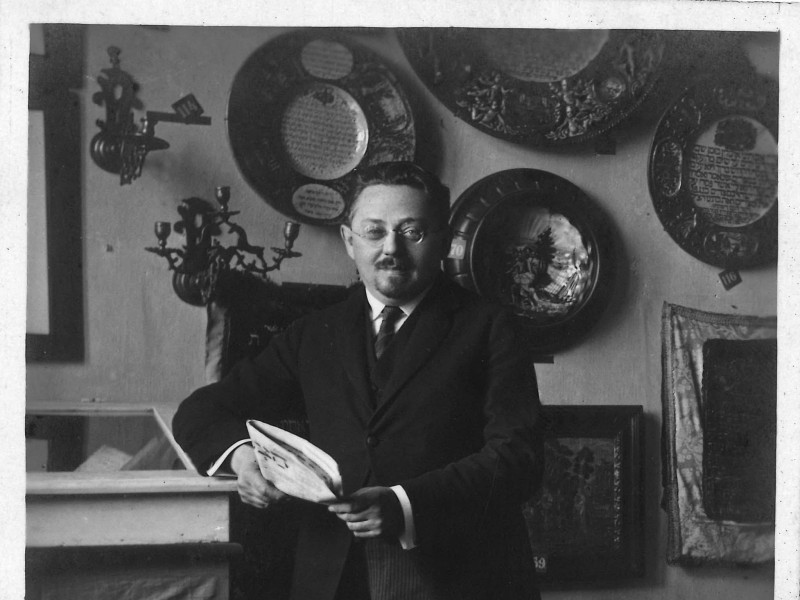 Ein Mann mit Brille und Anzug steht in einer Ausstellung und hält eine Zeitung in den Händen.