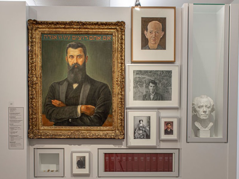 Ausstellungswand mit einem großen Porträt eines Mannes mit Bart und Anzug, umgeben von mehreren kleineren gerahmten Fotografien und Dokumenten an einer weißen Wand