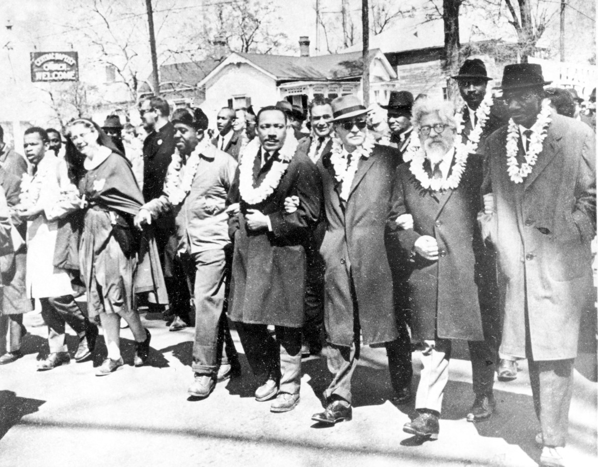 : Gruppe von Menschen in Mänteln und mit Blumenkronen, die Arm in Arm auf einer Straße stehen. Matin Luther King und Rabbi Heschel