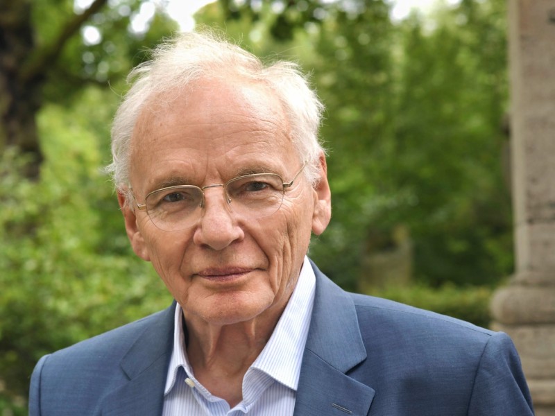 Ein älterer Mann mit grauen Haaren trägt einen blauen Anzug und ein hellblaues Hemd. Er steht vor einer grünen, bewaldeten Kulisse.