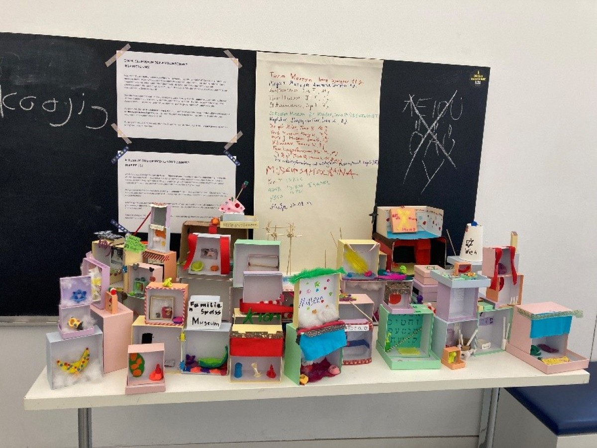 : Mehrere kleine, bunte Modellhäuser aus Pappe und Papier stehen auf einem weißen Tisch vor einer schwarzen Tafel mit handgeschriebenen Texten und Zeichnungen.