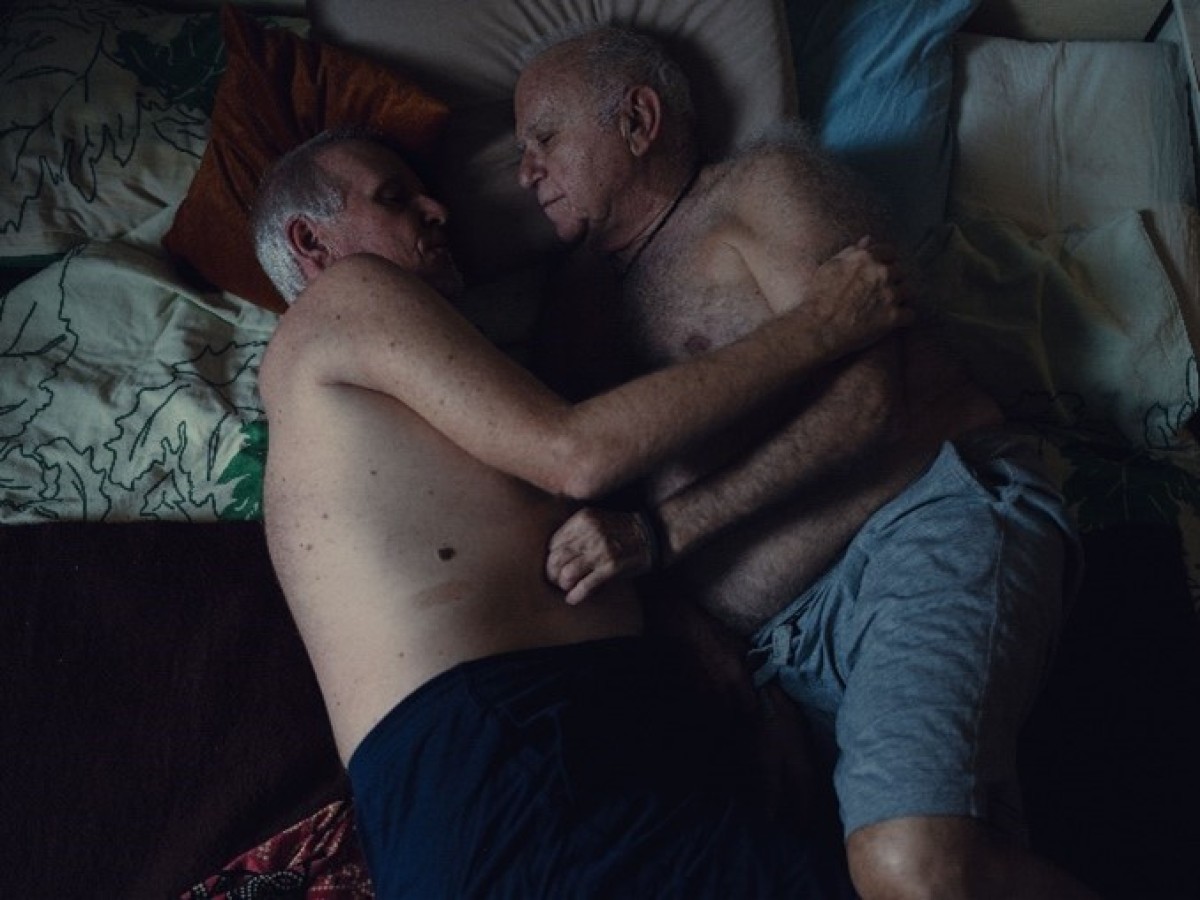 : Two elderly men lying in bed facing each other, embracing closely, one wearing dark shorts and the other light gray shorts