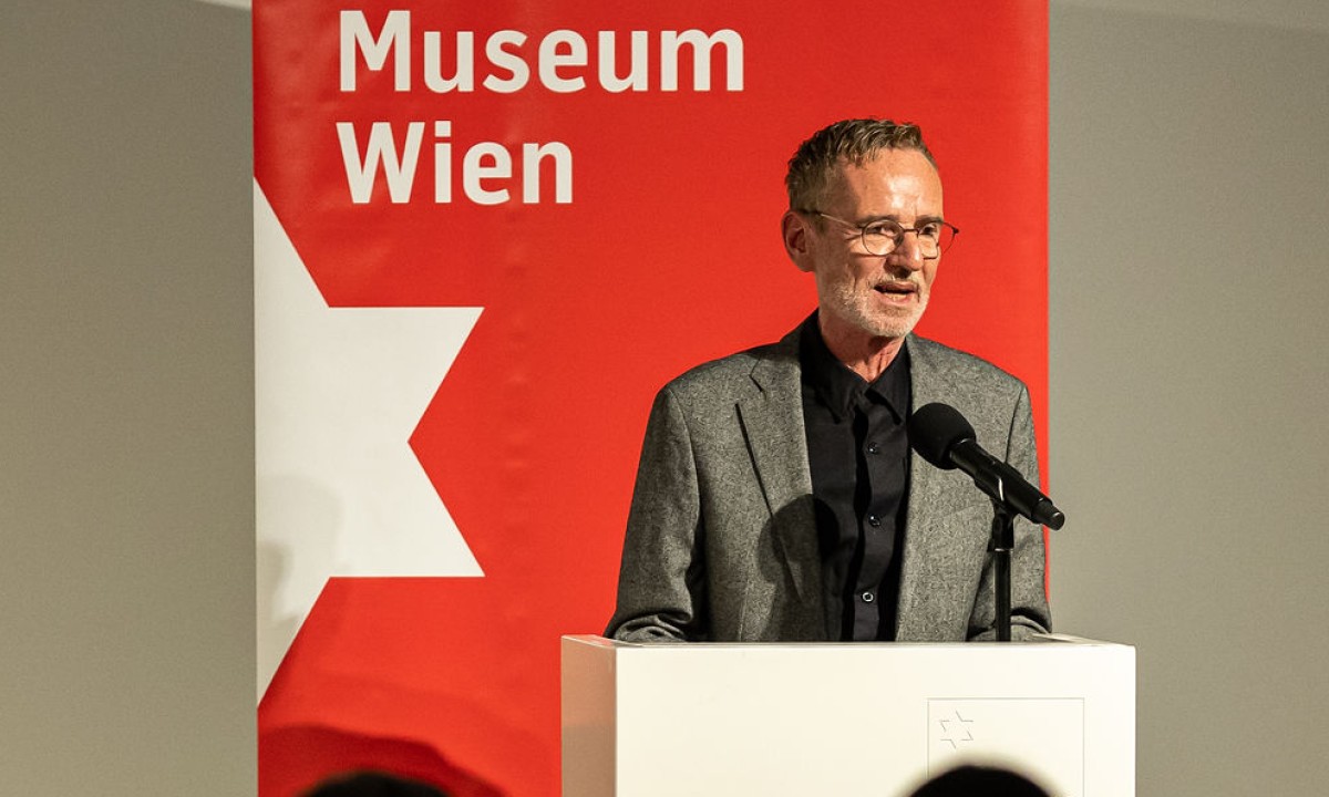 Mann steht am Rednerpult und hält eine Rede vor versammelten Besucher:innen.