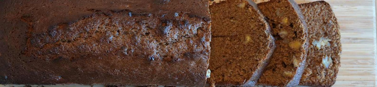 : Ein geschnittenes Stück Kuchen auf einem Holzbrett, mit mehreren Scheiben sichtbar. Der Kuchen hat eine braune Farbe und enthält Stücke von Nüssen oder Früchten.