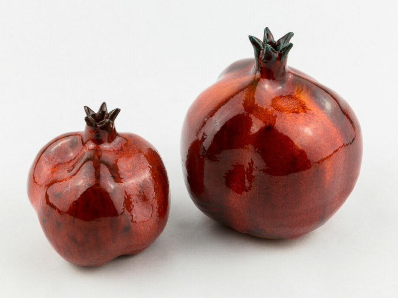 Zwei glänzende rote Granatäpfel aus Keramik, einer kleiner und einer größer, auf hellem Hintergrund