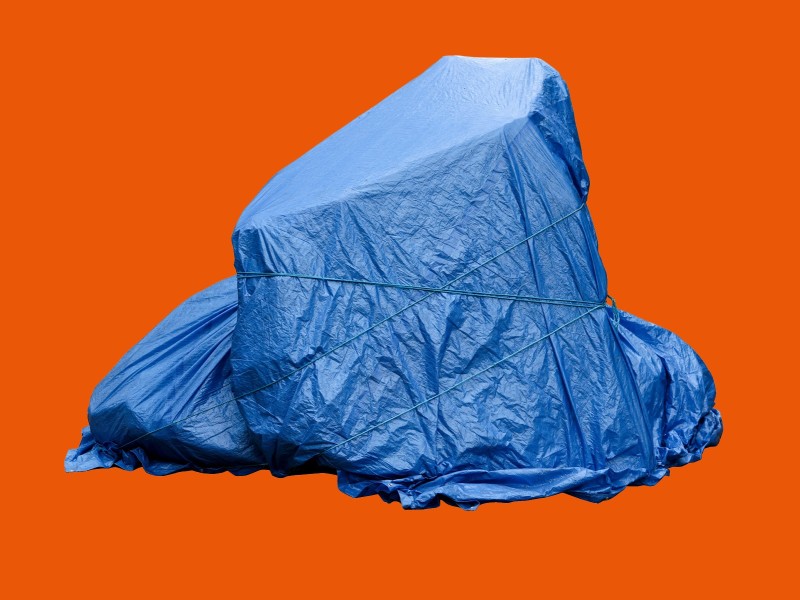 Großer Gegenstand mit blauer Plane abgedeckt und mit Seilen gesichert vor orangefarbenem Hintergrund