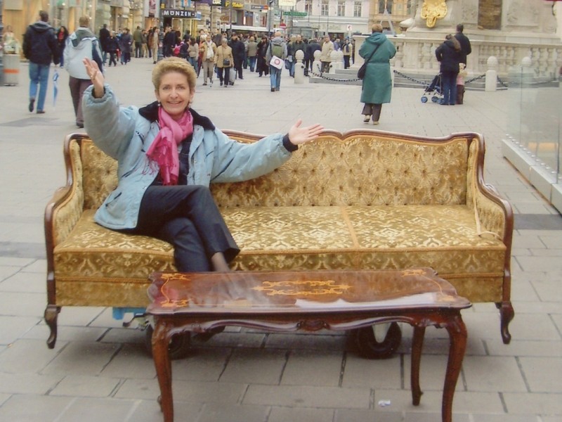 Frau mit Jeansjacke und pinkem Schal sitzt auf einem goldfarbenen Sofa auf einer belebten Fußgängerzone, umgeben von historischen Gebäuden.