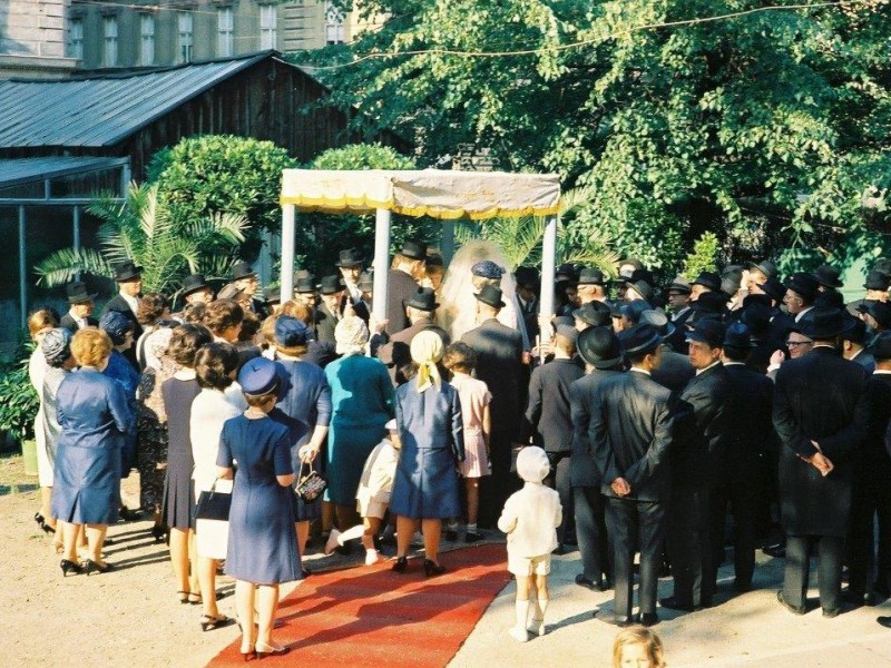 Group of people in formal attire gathered around a couple under a chuppah outdoors, surrounded by trees and buildings.