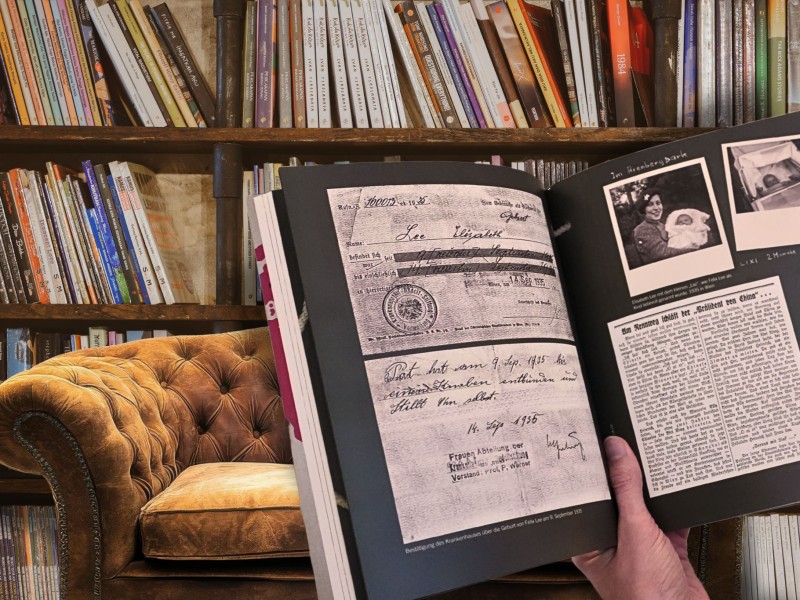 Person hält ein aufgeschlagenes Buch mit historischen Dokumenten und Fotos vor einem Bücherregal und einem braunen Samtsofa. KI Unterstützt