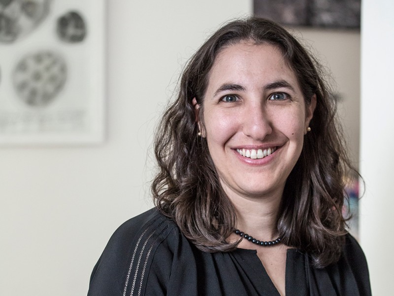 Woman with shoulder-length wavy hair wearing a black top holding a cup with both hands in front of a white wall with framed pictures.
