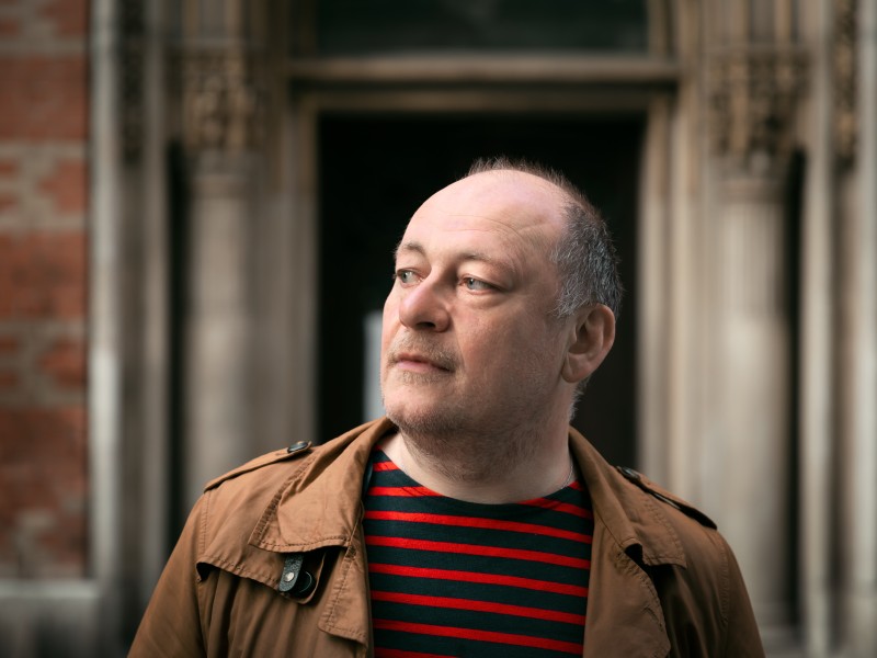 Person mit kurzem Haar, brauner Jacke und rot-schwarz gestreiftem Pullover vor einem historischen Gebäude mit Steinbogen.