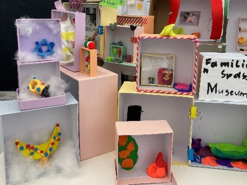 Mehrere kleine, bunte Dioramen aus Pappschachteln mit verschiedenen Figuren und Dekorationen, auf einem Tisch vor einer schwarzen Tafel und weißen Textblättern.