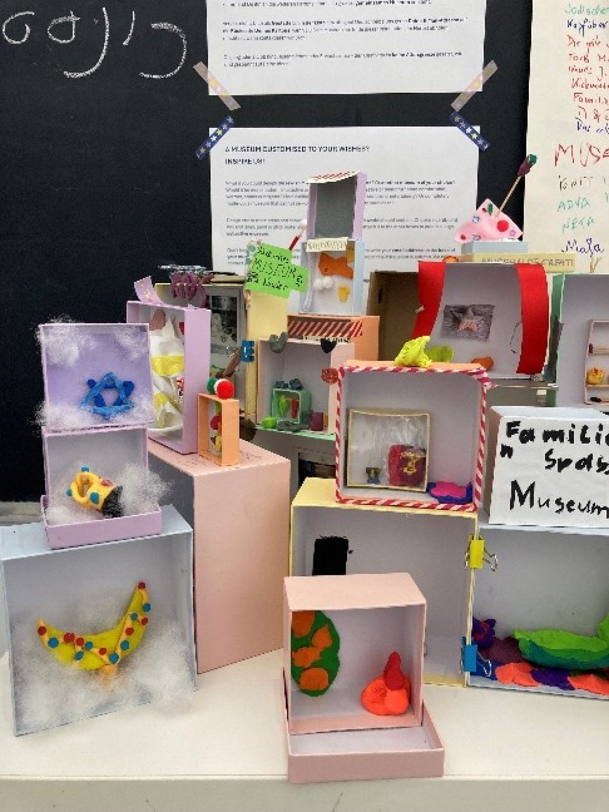 : Mehrere kleine, bunte Dioramen aus Pappschachteln mit verschiedenen Figuren und Dekorationen, auf einem Tisch vor einer schwarzen Tafel und weißen Textblättern.