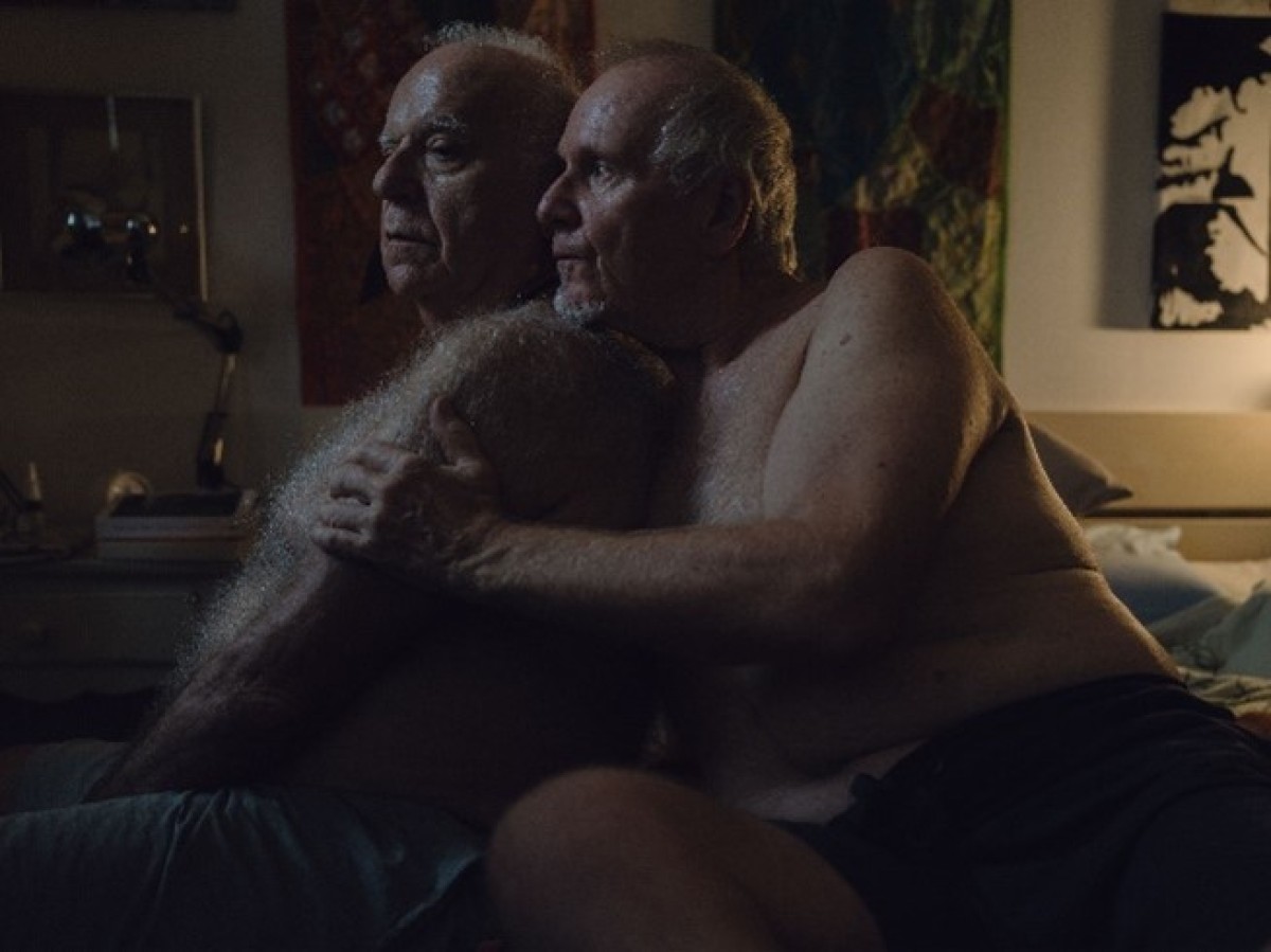 : Two elderly men sitting closely together in a dimly lit room, one embracing the other who rests their head on his shoulder