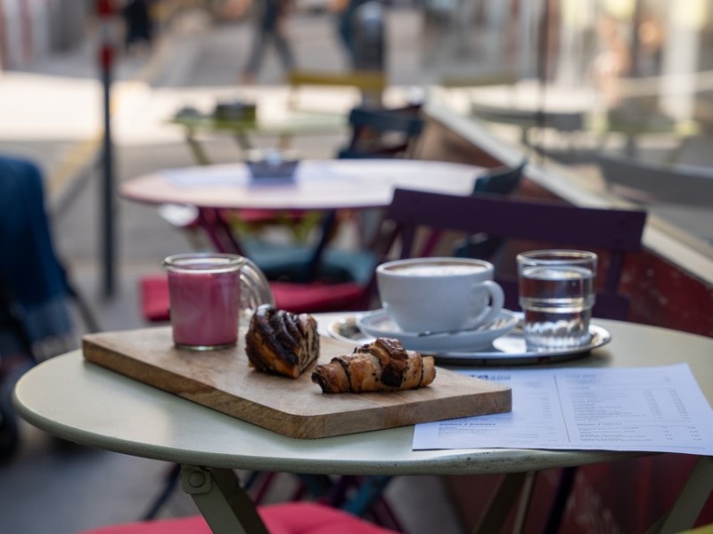Rundes Außencafé-Tisch mit zwei Croissants auf einem Holzbrett, einer Tasse Kaffee, einem Glas Wasser und einem rosa Kerzenglas, umgeben von farbigen Stühlen mit roten Sitzkissen