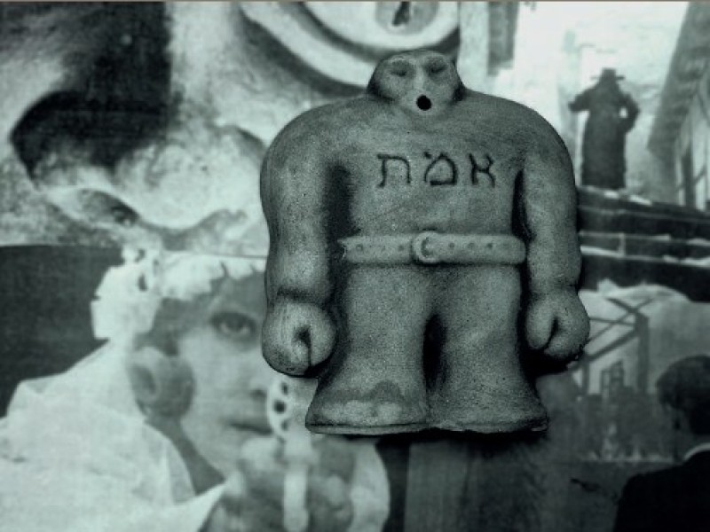 Skulptur einer stehenden Figur mit hebräischer Schrift auf der Brust, im Hintergrund unscharfe, überlagerte Gesichter und eine weitere Figur in einem urbanen Umfeld.