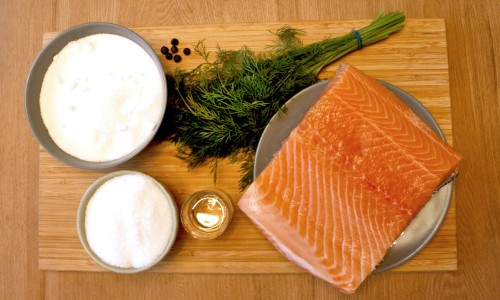 : Ein Holzbrett mit einem Stück Lachsfilet auf einem Teller, zwei Schüsseln mit Zucker und Salz, einem kleinen Glas mit Flüssigkeit, einem Bund Dill und einigen Wacholderbeeren.