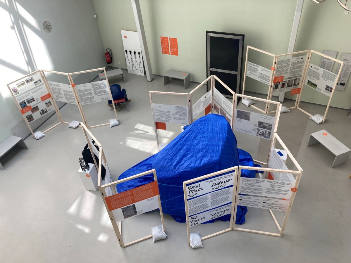 : Ausstellung mit Texttafeln auf Holzrahmen und einem Objekt unter einer blauen Plane in einem hellen Raum