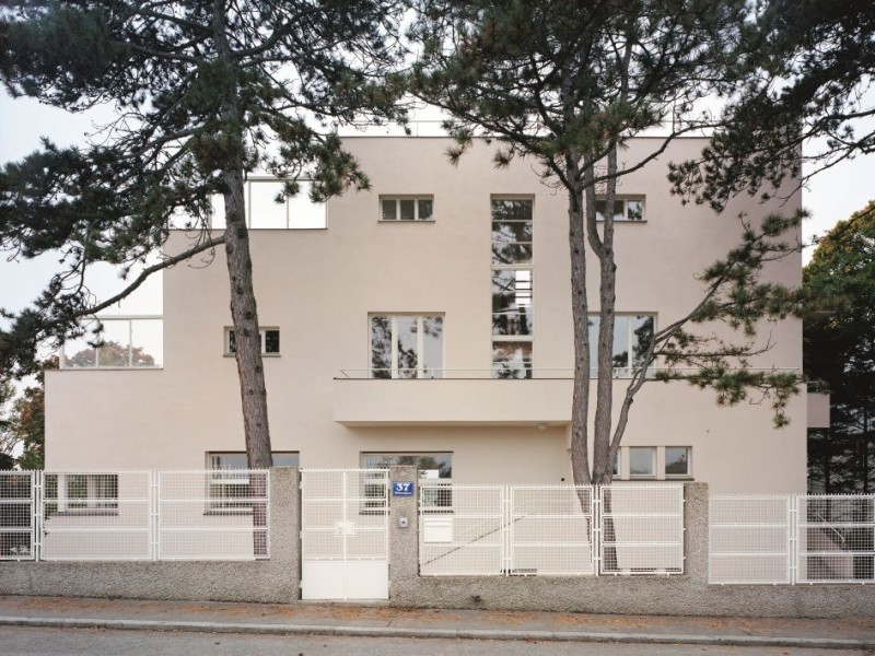 Kubisch geformtes Wohnhaus mit flacher Dachterrasse, umgeben von einem weißen Zaun und zwei hohen Bäumen im Vordergrund.