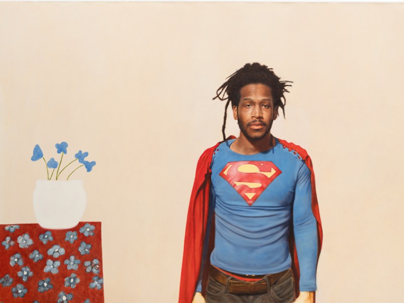 Mann mit Dreadlocks trägt ein blaues Superman-Shirt und einen roten Umhang, steht vor beige Hintergrund neben rotem Tisch mit weißer Vase und blauen Blumen