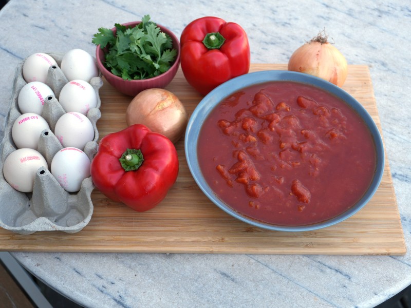 : Zwei rote Paprika, zwei Zwiebeln, ein Schälchen Koriander, ein Karton mit acht Eiern und eine Schüssel mit Tomatensauce auf einem Holzbrett.