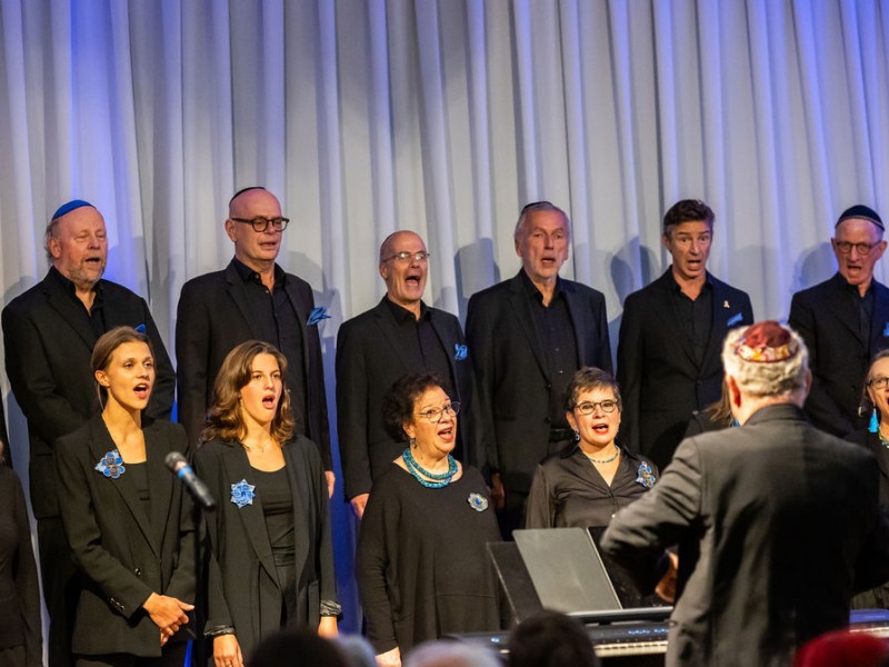 Choir with multiple people dressed in black in front of a light curtain, conducted by a person wearing a kippah and black suit