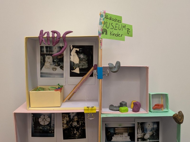 Kleines Diorama mit dem Wort "KIDS" aus lila Modelliermasse, zwei Fotos im Hintergrund und einem kleinen gelben Kasten mit grüner Deko im Vordergrund.