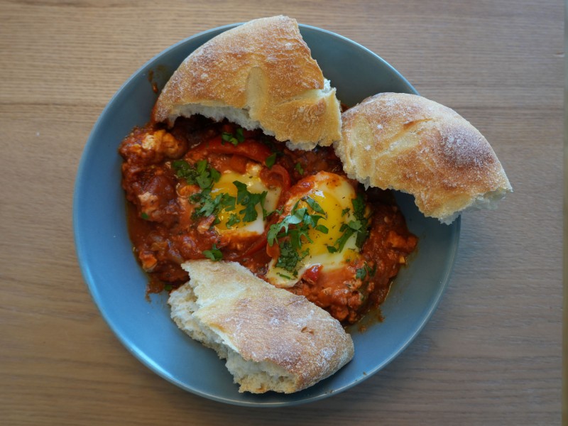 Blauer Teller mit zwei pochierten Eiern in Tomatensauce, garniert mit frischer Petersilie, dazu drei Stücke rustikales Weißbrot.