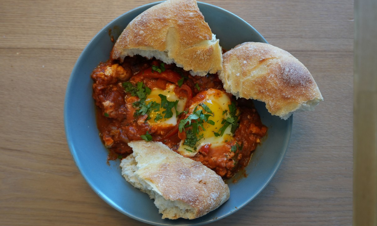 Blauer Teller mit zwei pochierten Eiern in Tomatensauce, garniert mit frischer Petersilie, dazu drei Stücke rustikales Weißbrot.