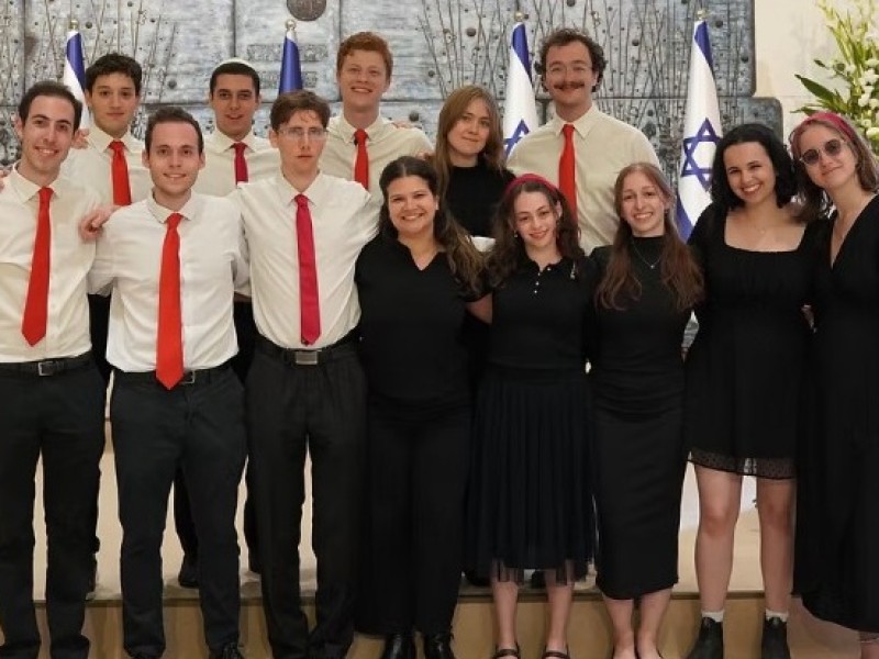 Group of people in formal attire standing in front of a wall with Israeli flags and floral arrangements.