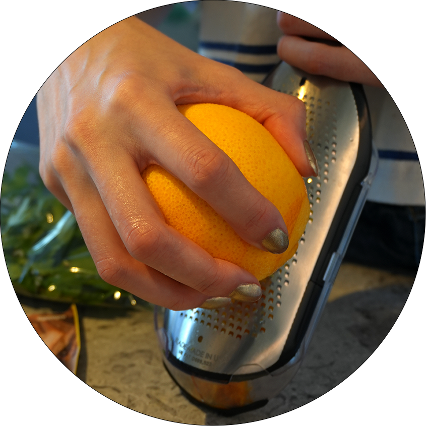 Eine Hand hält eine orangefarbene Zitrusfrucht über eine Reibe aus Metall. Die Reibe hat eine perforierte Oberfläche.
