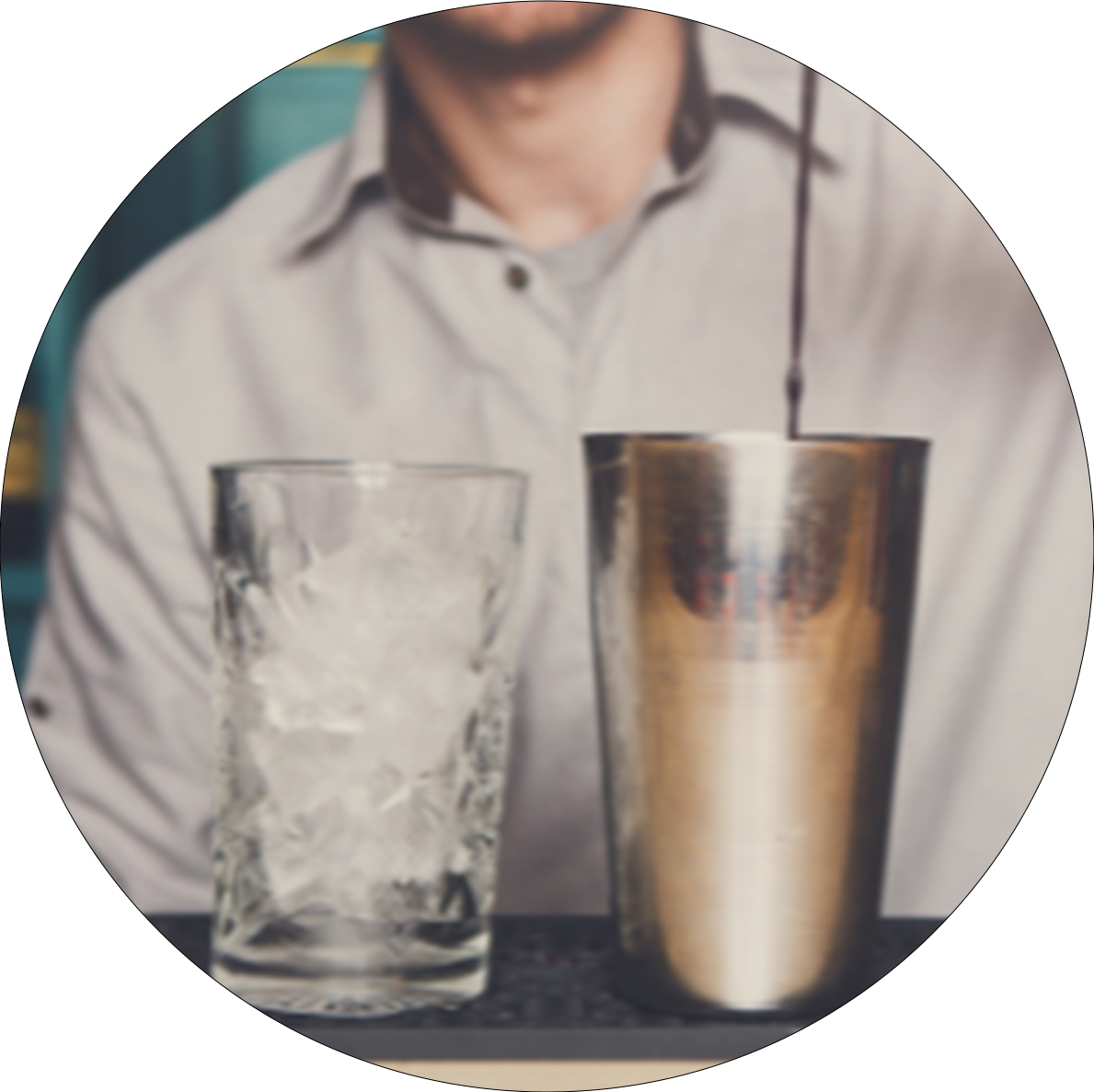 Zwei Gläser nebeneinander auf einer Theke, links ein mit Eis gefülltes Glas, rechts ein Metall-Shaker mit einem Rührstab