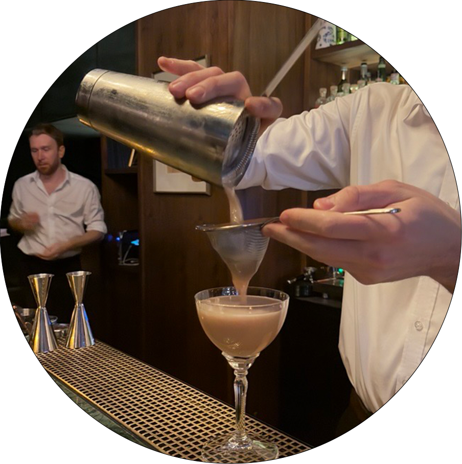 Eine Person gießt mit einem Barsieb einen cremigen Cocktail aus einem Metallshaker in ein Cocktailglas auf einer Bar.